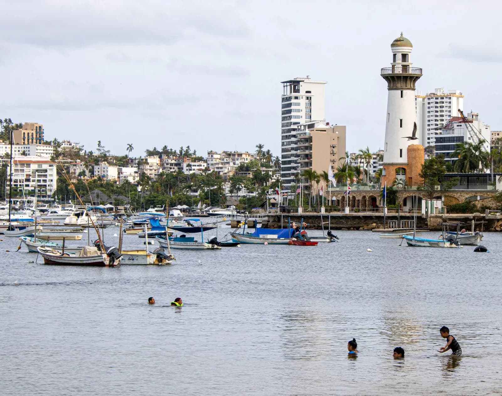 Bateaux amarrés et baigneurs au bord d'une plage de la baie d'Acapulco, avec un phare au loin