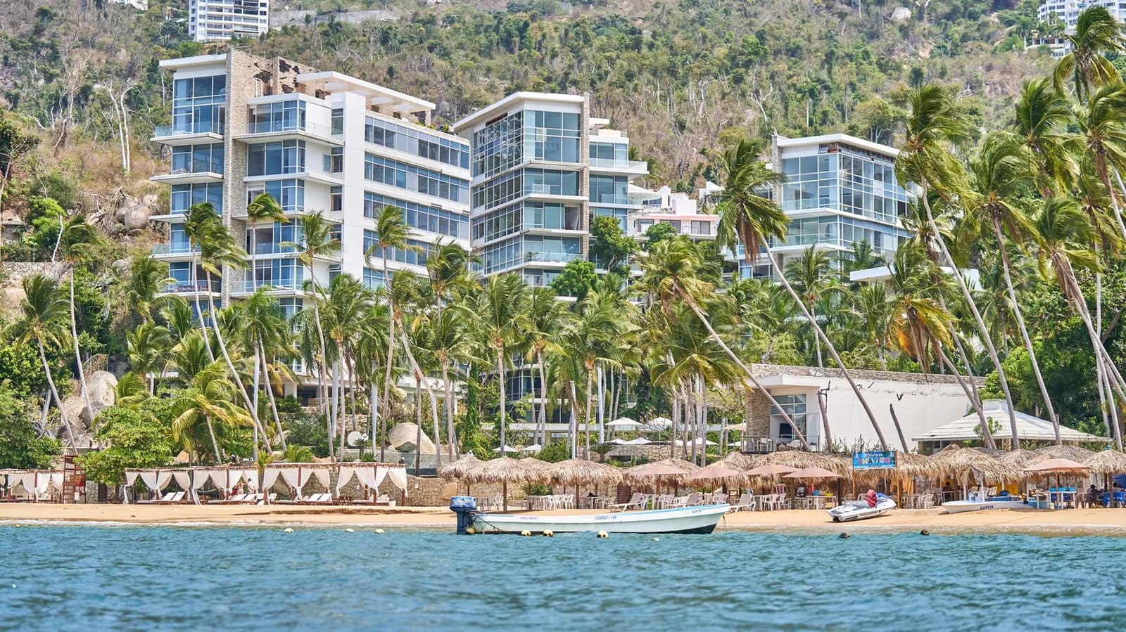 Plage de la baie d'Acapulco bordée de palmiers et de palapas, avec un bateau au bord de l'eau