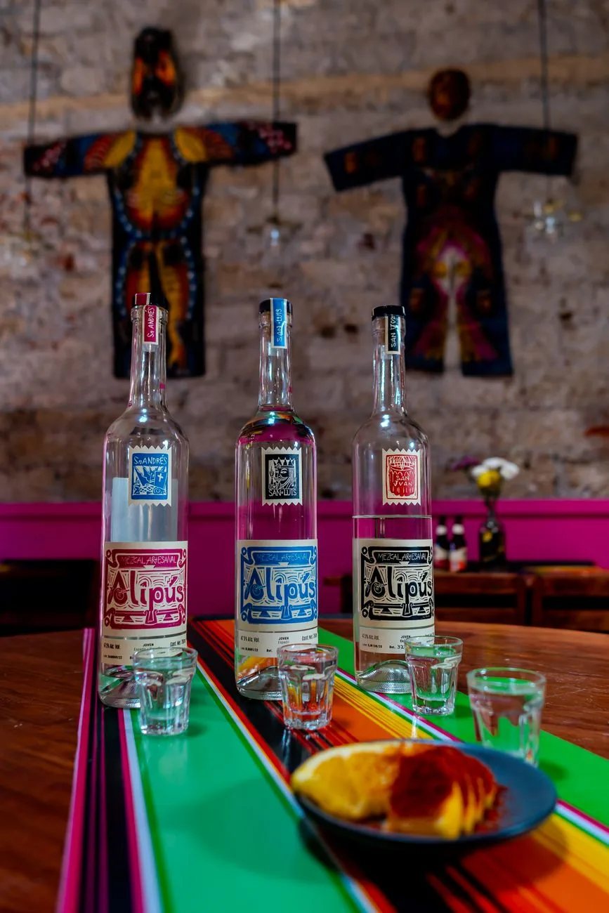 Trois bouteilles de mezcal artisanal alignées sur une table colorée dans un bar mexicain, avec verres et quartiers d'orange