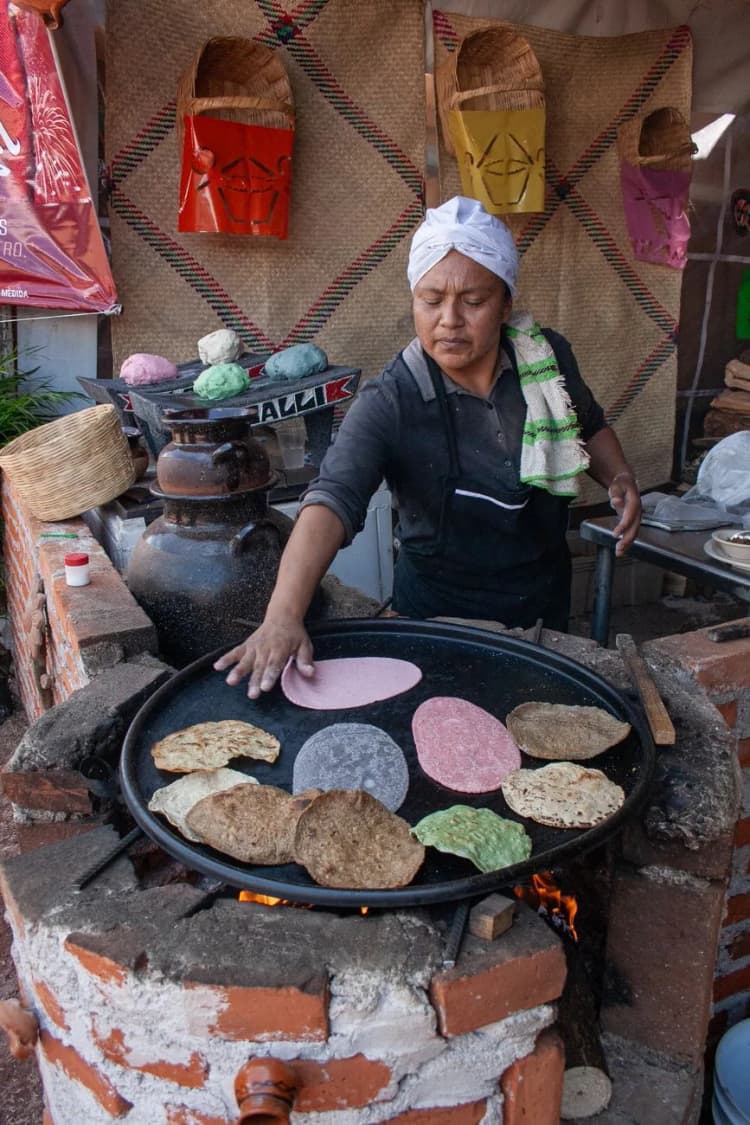 Cuisinière mexicaine faisant cuire des tortillas de maïs bleu, rose et vert sur un grand comal au feu de bois