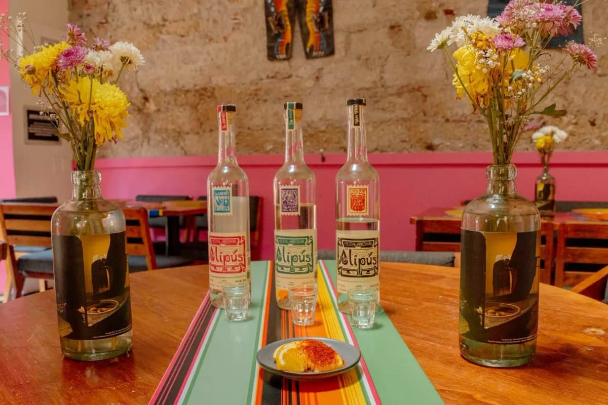 Trois bouteilles de mezcal artisanal aux étiquettes colorées sur une table en bois, avec une assiette d'orange