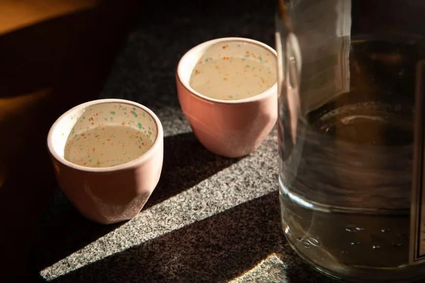 Deux petites coupes en terre cuite remplies de mezcal sur une dalle de pierre, bouteille en arrière-plan
