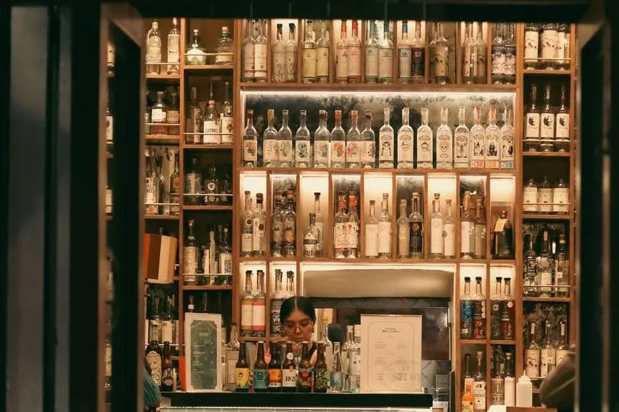 Mur d'étagères en bois rempli de bouteilles de mezcal derrière le comptoir d'un bar, éclairage chaud
